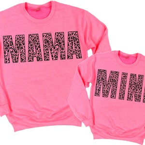 Könnte beinhalten: Zwei rosa Sweatshirts, eines groß und eines klein. Das größere Sweatshirt hat das Wort "MAMA" in schwarzen Blockbuchstaben mit Leopardenmuster. Das kleinere Sweatshirt hat das Wort "MINI" in schwarzen Blockbuchstaben mit Leopardenmuster. Beide Sweatshirts haben lange Ärmel.