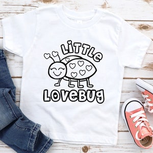 May include: White t-shirt featuring a black and white cartoon ladybug design with the text "Little Lovebug". The ladybug has heart-shaped spots and antennae. The shirt is paired with blue jeans and pink sneakers.