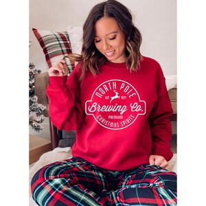 Może przedstawiać: Czerwona bluza z napisem "North Pole Brewing Co." w białym okrągłym wzorze. Bluza jest połączona ze spodniami od piżamy w kratę. Projekt zawiera renifera i słowa "Premium Christmas Spirits."
