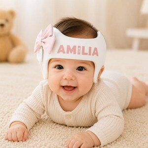 May include: A white cranial orthosis helmet featuring the name AMILIA in pink lettering on the front. The helmet is decorated with a light pink bow attached to the side. The design is smooth and contoured to fit the head, providing a clean appearance.