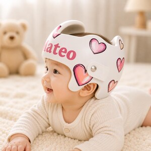 May include: A white cranial orthosis helmet featuring a pink heart pattern and the name Mateo printed in pink letters on the side. The helmet has a smooth, moulded shell with a metallic fastener on the side and cutouts around the ear area.