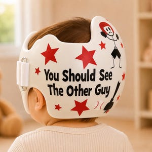 May include: A white cranial orthosis helmet featuring red star decals, a graphic of a stick figure with a sad expression, a baseball, and a baseball bat. The text "You Should See The Other Guy" is printed in black lettering on the side of the helmet.