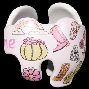May include: A white baby car seat with a whimsical design featuring a rainbow, cactus, flowers, cowboy boots, and a snake. The word "me" is written in pink on the side.