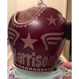 May include: Burgundy coloured helmet with a brown leather star and wings design. The helmet has a name "Harrison" written on the bottom.