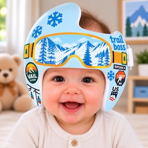 May include: A light blue cranial orthosis helmet featuring a graphic of ski goggles with a mountain landscape, blue snowflake icons, and various ski resort stickers including Vail, Loon, and the text trail boss printed on the side.