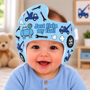 May include: A light blue cranial orthosis helmet featuring a pattern of dark blue tow trucks, black wrenches, tires, and white clouds. The text "Just fixin my flat!" is printed in dark blue letters across the front of the helmet, which has a white strap.