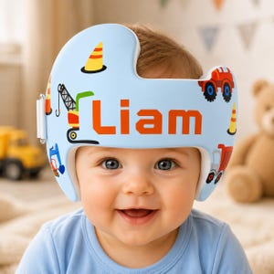 May include: A light blue cranial orthosis helmet featuring a construction-themed pattern with orange traffic cones, a crane, and trucks. The name Liam is printed in large orange block letters across the front. The helmet has a white side strap.