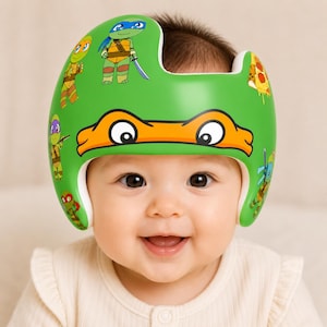 Könnte beinhalten: Ein leuchtend grüner Schädel-Orthesenhelm mit einem Grafikdruck von Teenage Mutant Ninja Turtles-Figuren und Pizzastücken. Die Vorderseite des Helms zeigt ein markantes orangefarbenes Augenmasken-Design mit schwarz-weißen Augen.