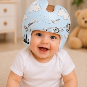 May include: A light blue protective cranial helmet featuring a black and white graphic print of clouds, paper airplanes, biplanes, and a city skyline. The helmet has a smooth, rounded shape with a cutout at the top and covers the sides of the head.