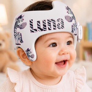 May include: A white cranial orthosis helmet featuring a repeating zebra pattern and the name Luna printed in black and white striped lettering on the front. The helmet has a smooth, moulded shell design with cutouts for the ears and a side adjustment dial.