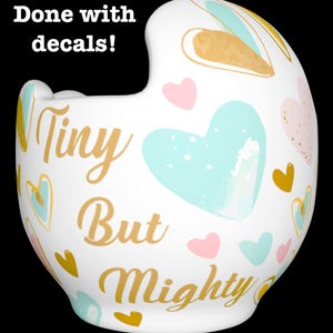 May include: White ceramic helmet with a gold inscription that reads "Tiny But Mighty" and a pattern of hearts in various colors, including pink, blue, and gold.