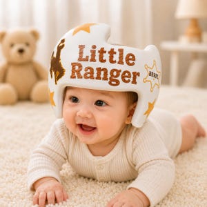 Könnte beinhalten: Ein weißer kranialer Orthesenhelm mit dem braunen Schriftzug Little Ranger, einer braunen Silhouette eines bockenden Pferdes, gelben Sternen und einer gelben Sheriffstern-Kontur mit dem Wort Brave auf der Seite des Helms.