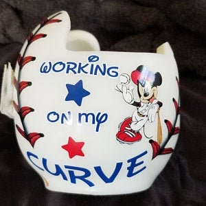 May include: White baseball helmet with a red, white, and blue Mickey Mouse design. The helmet has a baseball stitch pattern and the words "Working on my Curve" are written on the front.