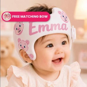 May include: A white protective cranial helmet featuring pink cartoon bear faces and the name Emma printed in pink script. The helmet includes a side adjustment dial and a pink graphic banner at the top with the text FREE MATCHING BOW.