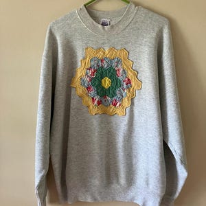 May include: A gray sweatshirt with a crew neck and long sleeves. The front features a colorful, floral-patterned appliqué in shades of yellow, green, pink, and blue. The sweatshirt is hanging on a green hanger.