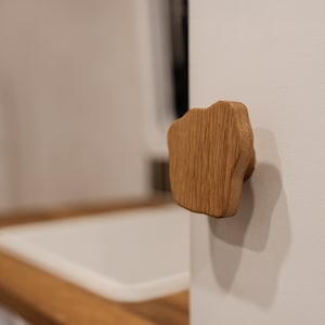 Può includere: Pomello per mobili in legno naturale dalla forma organica e irregolare. Il pomello è montato su una superficie bianca, mettendo in risalto le venature e i toni caldi del legno. Questo hardware unico aggiunge un tocco naturale.