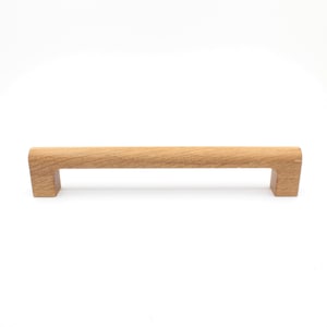 Puede incluir: Un tirador de armario de madera clara con una barra rectangular y extremos cuadrados. La veta de la madera es visible y el tirador está diseñado para un agarre fácil. Adecuado para cajones y armarios.