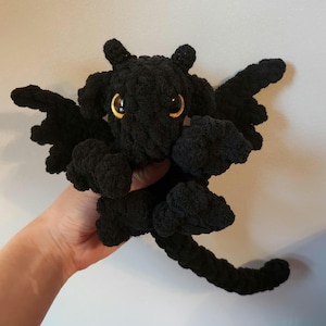 Puede incluir: Juguete de peluche de dragón negro de crochet con ojos amarillos. El dragón tiene alas y una cola larga.