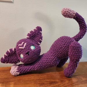 May include: A purple crochet cat toy in a stretching pose. The cat has light purple accents on its ears, paws, and tail tip. It has teal-colored eyes and white stitching details on its face. The toy is on a wooden surface.