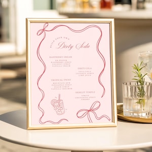 May include: A framed pink sign with the text "Build Your Own Dirty Soda" in a decorative font. The sign lists soda recipes like Raspberry Dream and Tropical Twist. A gold frame surrounds the sign, which is displayed on a white table with drinks in the background.