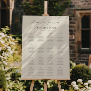 Może przedstawiać: Kremowy plan stołów weselnych na drewnianym sztaludze. Znak zawiera napis "please find your seat" w eleganckim czarnym skrypcie, wraz z imionami "Hannah & James" i datą "1 sierpnia 2025". Rozmieszczenie miejsc jest uporządkowane według numerów stołów.