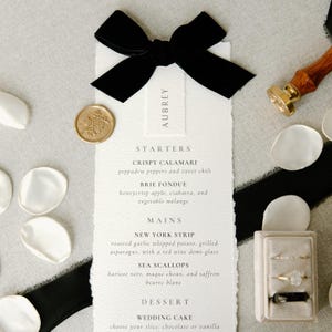 May include: A wedding menu with the name "AUBREY" printed on a white card, tied with a black velvet bow. The menu lists starters, mains, and dessert options. A gold wax seal and a ring box with rings are also visible.