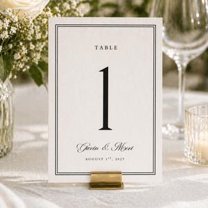 May include: A cream-colored table number card with a black border and the number 1. The card reads "TABLE" at the top, with the names "Gavin & Mori" and the date "August 1st, 2022" at the bottom. The card is held by a gold-colored metal clip.