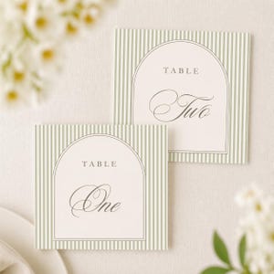 May include: Two square table place cards with a sage green and white striped background. Each card has a cream-colored arched shape with the word "TABLE" above the table number. One card reads "One" and the other reads "Two" in elegant script.
