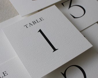 Op de afbeelding: Witte tafelnummerkaarten met zwarte cijfers en het woord 'TABLE' bovenaan gedrukt. De kaarten hebben een getextureerd oppervlak en zijn licht overlappend gerangschikt. Het nummer '1' is prominent aanwezig.