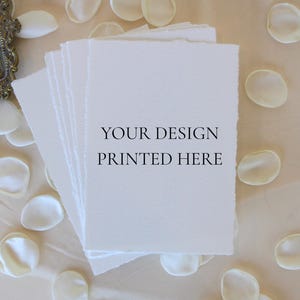Könnte beinhalten: Ein Stapel weißes strukturiertes Papier mit zerrissenen Kanten, mit dem Text "YOUR DESIGN PRINTED HERE". Das Papier ist von weißen Blütenblättern auf einer hellbeige Oberfläche umgeben. Eine dekorative Schale mit einem Wachssiegel ist sichtbar.