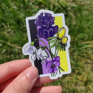 Può includere: Un adesivo con un design floreale con un'iride viola, fiori gialli, bucaneve bianchi e una viola viola. I fiori sono disposti su uno sfondo di strisce nere, viola e gialle, che rappresentano la bandiera non binaria.