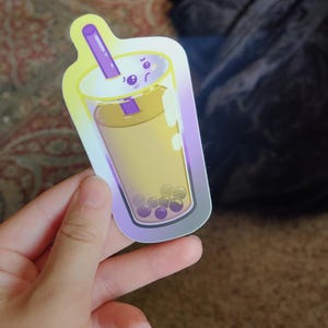 May include: A sticker featuring a colorful illustration of a bubble tea drink. The sticker has a gradient yellow and purple background, with a clear cup filled with a yellow liquid and dark purple boba pearls.