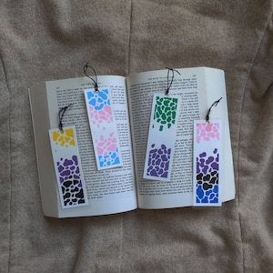Puede incluir: Cuatro marcapáginas coloridos con un patrón de manchas. Los marcapáginas son en tonos de rosa, verde, morado y azul. Los marcapáginas están hechos de papel y tienen una cuerda negra atada a la parte superior.