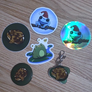 May include: Assortment of frog stickers, including a frog wearing a Santa hat, a frog on a lily pad, and a frog with a glittery background. The stickers are on a wooden surface.