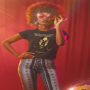 May include: A woman wearing a black t-shirt with a gold graphic of a face and the text "Now there she goes again... the dopest Ethiopian... The Pharcyde 92". She is wearing a pair of pink sunglasses and has an afro hairstyle.