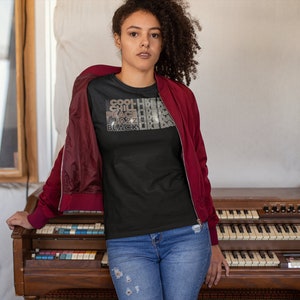 May include: A woman wearing a black t-shirt with the text "COOL LIKE A CHILL LIKE A PEACE LIKE A JAZZ LIKE A BLACK LIKE DA" in gold glitter lettering. She is standing in front of a vintage organ.
