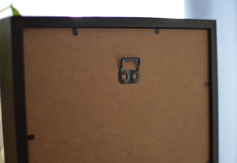 May include: Back of a black picture frame with a brown backing. A black metal hanger is centered on the frame, with two holes for hanging. The frame is designed for wall display.