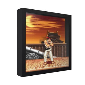May include: Framed pixel art of a Street Fighter character in a fighting stance. The artwork features a sunset background with a cannon and a traditional Japanese building. The character is wearing a white gi with a red headband.