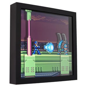May include: A framed 3D shadow box featuring a pixelated scene from a video game. The artwork depicts a blue character shooting a blue energy blast at a robot. The frame is black.