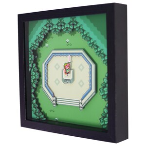 May include: A framed 3D art piece featuring a pixelated scene from a video game. The artwork depicts a character holding a glowing sword in a green landscape with a white and blue structure. The frame is black.
