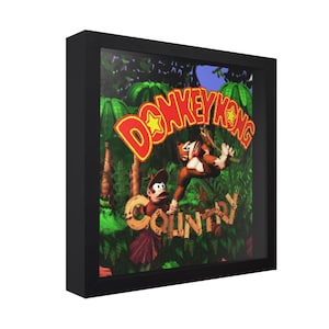 May include: A framed print featuring the Donkey Kong Country video game logo and characters. The artwork shows Donkey Kong and Diddy Kong in a jungle setting, with the game's title in bold, colorful lettering. The frame is black.