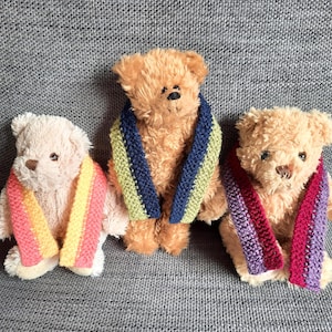 May include: Three plush teddy bears with colorful knitted scarves. The bears are light brown and cream-colored, each wearing a different scarf with stripes of pink, yellow, blue, green, and purple. The bears are sitting on a gray surface.