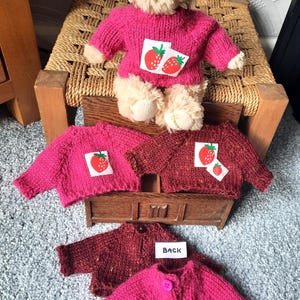 May include: A teddy bear in a pink strawberry sweater, with several miniature pink and maroon sweaters, some with strawberry patches. The word "Back" is visible on a small white tag. The sweaters are hand-knitted.