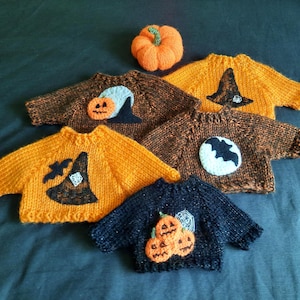 Op de afbeelding: Een collectie van vijf kleine, handgebreide truien in oranje, bruin en zwart, voorzien van Halloween-thema applicaties zoals pompoenen, heksenhoeden, vleermuizen en een spinnenweb, vergezeld door een enkele kleine, gebreide oranje pompoen.