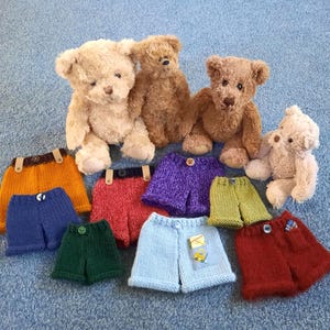 Könnte beinhalten: Vier Plüsch-Teddybären in verschiedenen Braun- und Beigetönen sind hinter einer Sammlung von neun kleinen, handgestrickten Shorts in den Farben Orange, Blau, Rot, Lila, Grün und Hellblau angeordnet, einige davon mit Knöpfen.