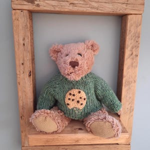 Op de afbeelding: Een pluche teddybeer met een roze kop en poten, gekleed in een groene trui met een koekjesontwerp. De beer zit op een houten plank in een rustieke houten lijst. De achtergrond is een lichtblauwe muur.