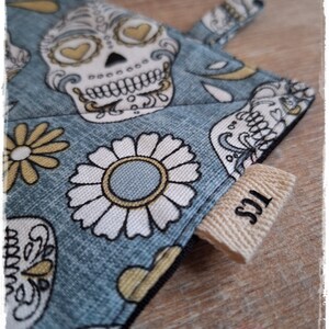 Könnte beinhalten: Blauer Stoff-Topflappen mit einem weißen und gelben Zucker-Schädel-Design und einem weißen Gänseblümchen-Muster. Der Topflappen hat ein braunes Stoff-Etikett mit dem Text "123" darauf.