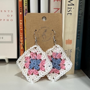 May include: A pair of square crochet earrings featuring a granny square pattern in white, pink, and blue yarn. The earrings are attached to a brown cardboard display card with the text Sunny Side Store Co. Cincinnati and a hand illustration.