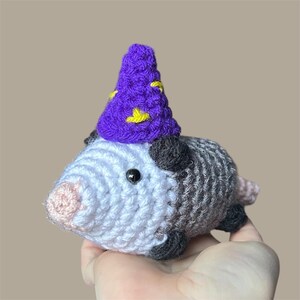 May include: A crocheted opossum figurine with a purple wizard hat adorned with yellow stars. The opossum has a gray and white body, black ears, and a pink nose. The figurine is held in a hand.
