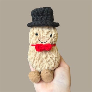 May include: A plush peanut toy with a black top hat and a red bow tie. The peanut has a smiling face and corduroy feet. The toy is beige and brown.